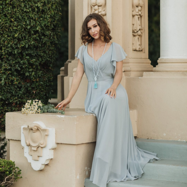 Woman in a logn blue flowy dress with a pearl necklace sitting on the steps of a colosseum.